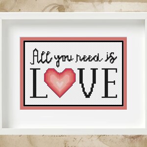 Może przedstawiać: Haftowany krzyżykiem obraz w białej ramie z czerwonym obramowaniem. Haftowany obraz przedstawia napis "All you need is LOVE" z różowym sercem w środku.