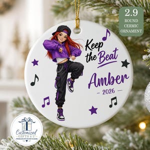 May include: A round white ceramic ornament featuring a cartoon character with red hair wearing a black cap, purple hoodie, and black cargo pants. The text reads Keep the Beat, Amber, and 2026, surrounded by purple and black musical notes and stars.