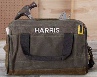 Puede incluir: Una bolsa de herramientas de lona verde con el nombre HARRIS bordado en la parte delantera. La bolsa tiene un asa negra y un cierre de cremallera. Está llena de herramientas, incluyendo un martillo, alicates y una navaja.