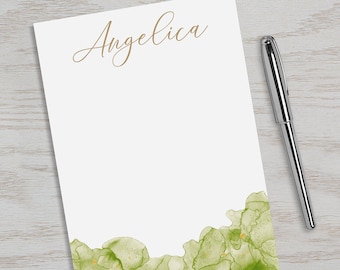 以下が含まれることがあります： 上部に金色の文字で「Angelica」と書かれた白いメモ帳。メモ帳の下部には、金色のアクセントがついた緑色の水彩画のデザインが施されています。