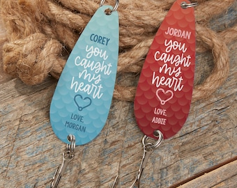 Pode incluir: Duas amostras de pesca personalizadas. Uma é azul, a outra vermelha, ambas com o texto "you caught my heart". A amostra azul diz "Corey" e "Love, Morgan", a vermelha diz "Jordan" e "Love, Addie". Ambas têm um anzol triplo.