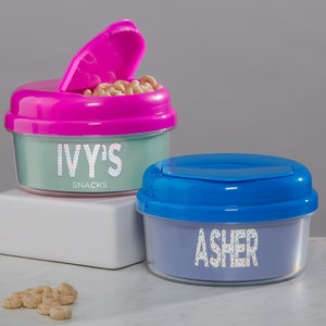 May include: Two plastic snack containers with lids. The container on the left is teal with a pink lid and says "IVY'S SNACKS". The container on the right is blue with a blue lid and says "ASHER".