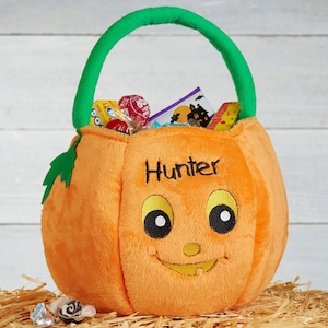May include: Orange plush pumpkin-shaped Halloween candy basket with a green handle and leaf accents. The basket has a smiling face and the name "Hunter" embroidered on the front. The basket is filled with various candies.