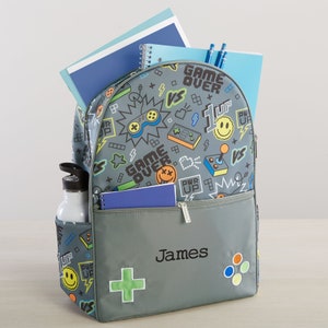 May include: A gray backpack with a colorful video game design. The backpack has a large front pocket with a zipper and a smaller pocket on the side. The backpack is personalized with the name "James".