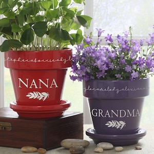 May include: Two terracotta flower pots, one red and one purple, with white lettering. The red pot says "NANA" and the purple pot says "GRANDMA". Both pots have a white leafy design.