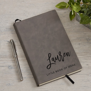 May include: A gray faux leather journal with a black ribbon bookmark. The journal has the name "Lauren" and the text "Little Book of Ideas" printed on the front cover.