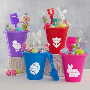 May include: Four colorful plastic buckets filled with Easter treats and toys. Each bucket has a different name on it: Taylor, Sandra, Christopher, and Abigail. The buckets are sitting on a wicker basket.