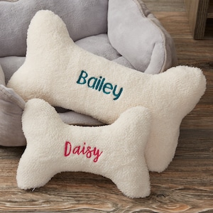 May include: Two cream-colored, bone-shaped dog pillows. The larger pillow has the name "Bailey" embroidered in teal, and the smaller pillow has "Daisy" embroidered in pink. The pillows are placed in front of a gray dog bed.