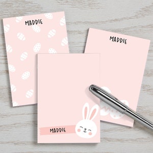 May include: Three pink notepad sets with the name Maddie on each. One notepad has a bunny with a pink ribbon and the text Maddie on it. Another notepad has a pink background with white eggs. The third notepad is plain pink.
