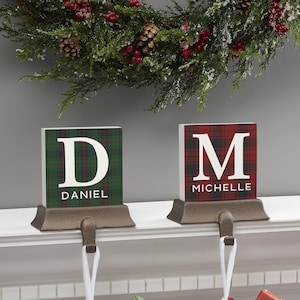 May include: Two square stocking holders with bronze-toned metal bases and hooks. The left holder features a green plaid pattern with a white letter D and the name DANIEL. The right holder features a red plaid pattern with a white letter M and the name MICHELLE.