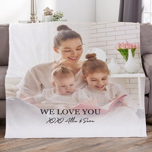 May include: White fleece blanket featuring a family portrait of a mother and two children reading a book. The text "WE LOVE YOU" and names are printed at the bottom. The blanket is draped over a gray sofa.