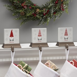 May include: Three Christmas stocking holders with gnome designs. Each holder has a white block with a gnome illustration and a name. The gnomes wear red hats and gray beards. The holders are on a white mantel with white stockings.