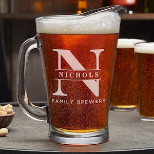 May include: A clear glass beer pitcher with a handle, filled with a dark amber beer. The pitcher is etched with the letter "N" and the text "Nichols Family Brewery".