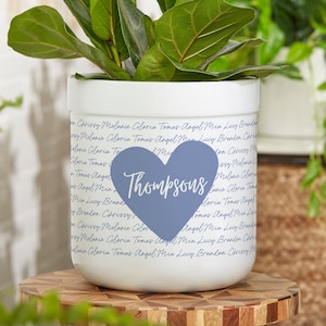 May include: White ceramic planter with a blue heart-shaped decal that reads "Thompsons." The planter is filled with a green plant. The planter's exterior is covered with names in a script font.