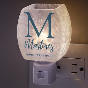 May include: A white night light with a frosted glass shade. The shade has a blue "M" and the name "Martinez" written in cursive. The text "Home Sweet Home" is also written on the shade. The night light is plugged into an outlet.