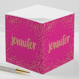 May include: A hot pink sticky note cube with the name "Jennifer" in gold script. The cube is decorated with gold and pink confetti. A pen with a crystal-studded barrel and gold accents rests beside it.