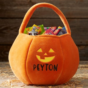 May include: Orange plush pumpkin-shaped Halloween candy basket with a handle. The basket features a jack-o'-lantern face and the name "Peyton" embroidered in black. The basket is filled with various wrapped candies.