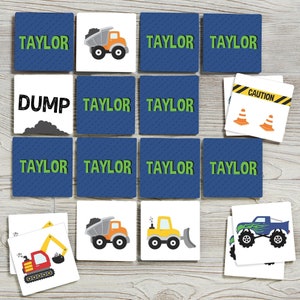 May include: A set of 12 blue and white square cards with images of construction vehicles and the word "Taylor" on some of the cards. The cards are arranged in a grid on a wooden surface. The cards include images of a dump truck, a bulldozer, an excavator, and construction cones.