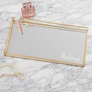 May include: A gold-framed mirrored tray with the name Mackenzie etched on the surface. The tray is on a white marble surface.