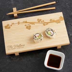 May include: A bamboo serving tray with a floral design and the text "The Wallaces Soy Happy Together". Two sushi rolls are on the tray, and a small white bowl with soy sauce is in the foreground.