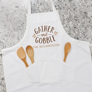 May include: White kitchen apron with a brown print that says "Gather and Gobble" with the name "The Richardsons" underneath. The apron has a large pocket and two wooden spoons are in the pocket.