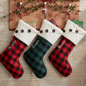 May include: Three personalized Christmas stockings with white faux fur cuffs and button accents. The stockings are red and black buffalo plaid, green and black buffalo plaid, and red and black buffalo plaid. The names Kaitlyn, Connor, and Morgan are embroidered on the stockings.