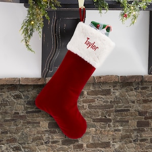 May include: A deep red velvet Christmas stocking featuring a plush white faux fur cuff. The name Taylor is embroidered in red script on the white cuff. A small gift wrapped in green paper with a white ribbon is tucked inside the stocking.