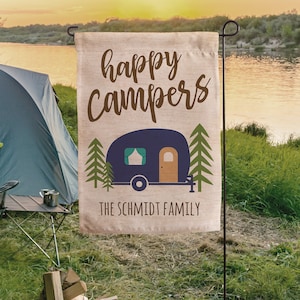 May include: A beige garden flag with the text "happy campers" in brown lettering. A blue camper with a brown door and green trees is pictured on the flag. The text "THE SCHMIDT FAMILY" is at the bottom of the flag.