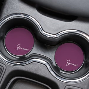 May include: Two round car coasters in a car cup holder. The coasters are a deep purple color with the name "Brianna" in white script. The cup holder is black.