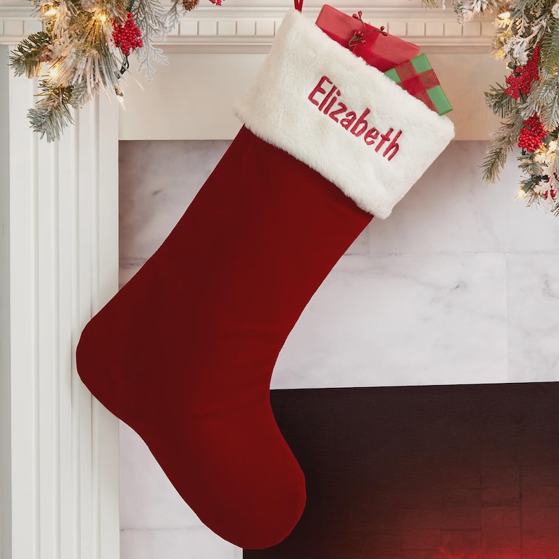 May include: A deep red velvet Christmas stocking featuring a plush white faux fur cuff. The name Elizabeth is embroidered in red script on the white cuff. A small gift box wrapped in red and green paper is tucked inside the top of the stocking.