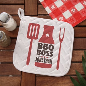 May include: White quilted oven mitt with a red BBQ design. The design includes a spatula, fork, and a bottle with the words "BBQ BOSS JONATHAN". A red and white checkered napkin is in the background.