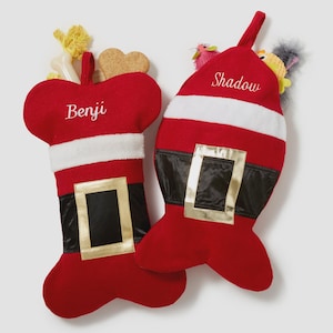 May include: Two red holiday stockings shaped like a dog bone and a fish. The bone stocking features the name Benji and a white faux fur trim with a black belt and gold buckle. The fish stocking features the name Shadow with similar belt details.
