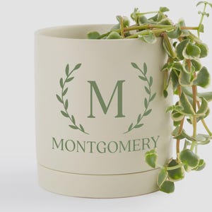May include: A cream-colored cylindrical planter with a green laurel wreath design, the letter "M", and the name "Montgomery". Trailing green and white variegated plant stems cascade from the top of the planter.