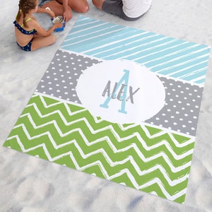 May include: A personalized beach blanket with a green chevron pattern, a gray polka dot pattern, and a blue striped pattern. The blanket features a white circle with the name "Alex" in blue lettering.