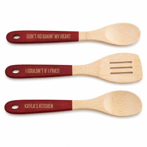 May include: Three wooden kitchen utensils with light tan heads and dark red handles. The top spoon features the text DON'T GO BAKIN' MY HEART, the middle slotted turner says I COULDN'T IF I FRIED, and the bottom spoon is engraved with KAYLA'S KITCHEN.