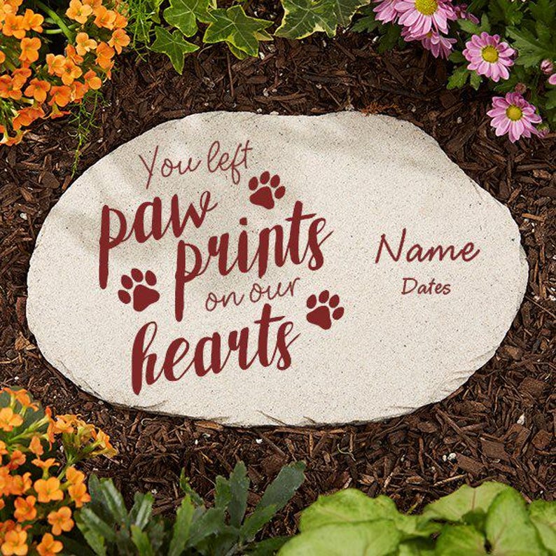 May include: A light-colored stone garden marker with the words "You left paw prints on our hearts" in burgundy script. Paw print icons are also included. Space is available for a name and dates.
