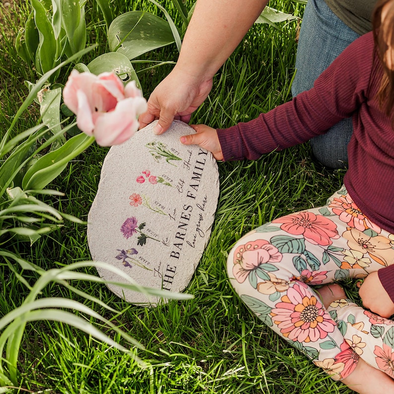 Pu&ograve; includere: Un segnaposto da giardino in pietra grigia con illustrazioni floreali e la scritta "THE BARNES FAMILY love grows here". Il segnaposto &egrave; tenuto da due persone in un ambiente esterno erboso. Un tulipano rosa &egrave; sullo sfondo.