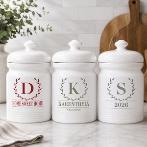 May include: Three white ceramic canisters with lids, each featuring a different design. One has a red "D" with "HOME SWEET HOME", another has a green "K" with "KARENTHYIA WELCOME!", and the last has a gray "S" with "2026".