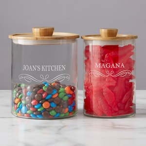 May include: Two glass canisters with bamboo lids. The canister on the left has colorful candies inside and is labeled "JOAN'S KITCHEN". The canister on the right has red fish-shaped candies inside and is labeled "MAGANA".