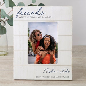 K&ouml;nnte beinhalten: Wei&szlig;er Holzbilderrahmen mit einem wei&szlig;en Passepartout und einem Foto von zwei Freundinnen. Der Rahmen tr&auml;gt den Text "friends are the family we choose" und "Sasha + Jodi best friends, wild adventures!" aufgedruckt.