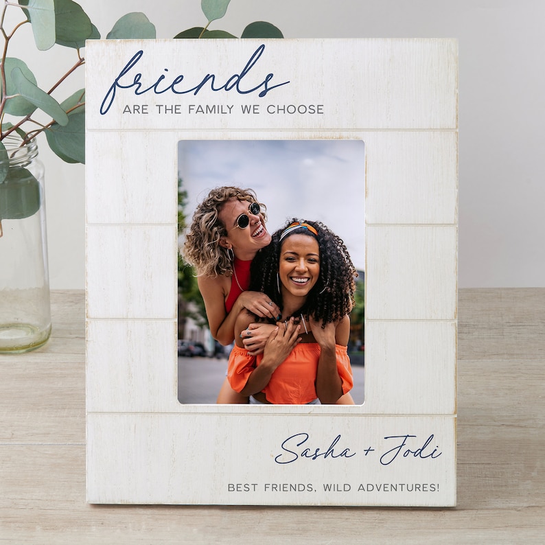 K&ouml;nnte beinhalten: Wei&szlig;er Holzbilderrahmen mit einem wei&szlig;en Passepartout und einem Foto von zwei Freundinnen. Der Rahmen tr&auml;gt den Text "friends are the family we choose" und "Sasha + Jodi best friends, wild adventures!" aufgedruckt.