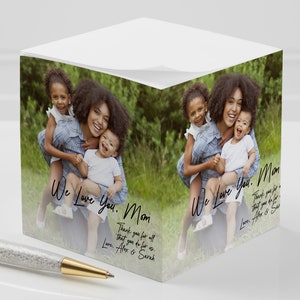 May include: A white cube-shaped notepad with a photo of a mother and her two children. The photo is surrounded by a green and brown blurred background. The text "We Love You. Mom" is written in black cursive on the notepad. The text "Thank you for all that you do for us. Love. Alex & Sarah" is written in black cursive on the notepad.