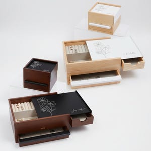 May include: Wooden jewelry boxes in various sizes and colors. Some boxes are light wood, others are dark brown. Each box has a drawer and a lid with a floral design. The lids are engraved with names like "Jennifer" and "Charlotte".