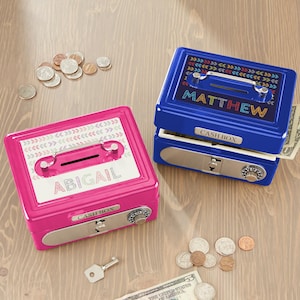 May include: Two personalized cash boxes, one pink and one blue, sit on a wooden surface. The pink box has "ABIGAIL" printed on it, while the blue box says "MATTHEW". Both boxes have a combination lock, a slot for money, and a handle. Coins and a key are also visible.