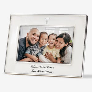 May include: A rectangular silver-toned picture frame with a glossy white border. The top center features a small silver cross. The bottom border is inscribed with the black text "Bless This Home" and "The Hamiltons" in a cursive script font.