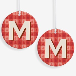 May include: Two round Christmas ornaments with the letter "M". The ornaments have a red and white plaid pattern with small Christmas trees. The letter "M" is in a cream color with a brown outline. Each ornament has a white string for hanging.