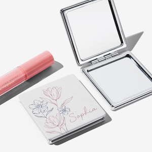 May include: A silver compact mirror is open, reflecting the light. A white square compact with a floral design and the name "Sophia" is next to it. A pink lip gloss tube is also in the image.