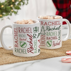 May include: Two white ceramic mugs with a Christmas-themed design. The mugs feature the name "Rachel" repeated in a variety of fonts and sizes, along with images of a Christmas tree, a gift box, and an ornament. The mugs are filled with hot chocolate and marshmallows.