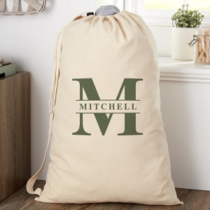 May include: A large, cream-colored laundry bag with a drawstring closure. The bag features a large, dark green letter "M" with the name "MITCHELL" printed below it. The bag has a shoulder strap.
