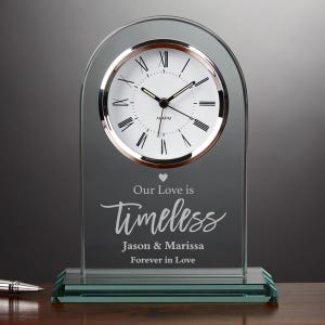 Op de afbeelding: Een heldere glazen klok met een zilverkleurige rand en Romeinse cijfers. De wijzerplaat leest "ALARM". Daaronder staat de tekst "Our Love is Timeless" en "Jason & Marissa Forever in Love".